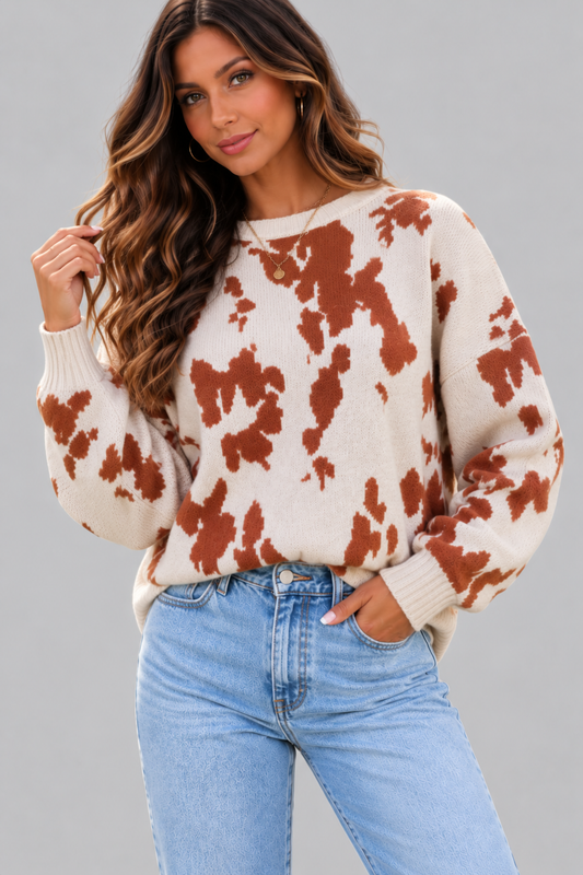 A woman wearing a cream-colored knit sweater with a rust-brown tie-dye pattern, paired with light-wash blue jeans, against a solid gray studio background.