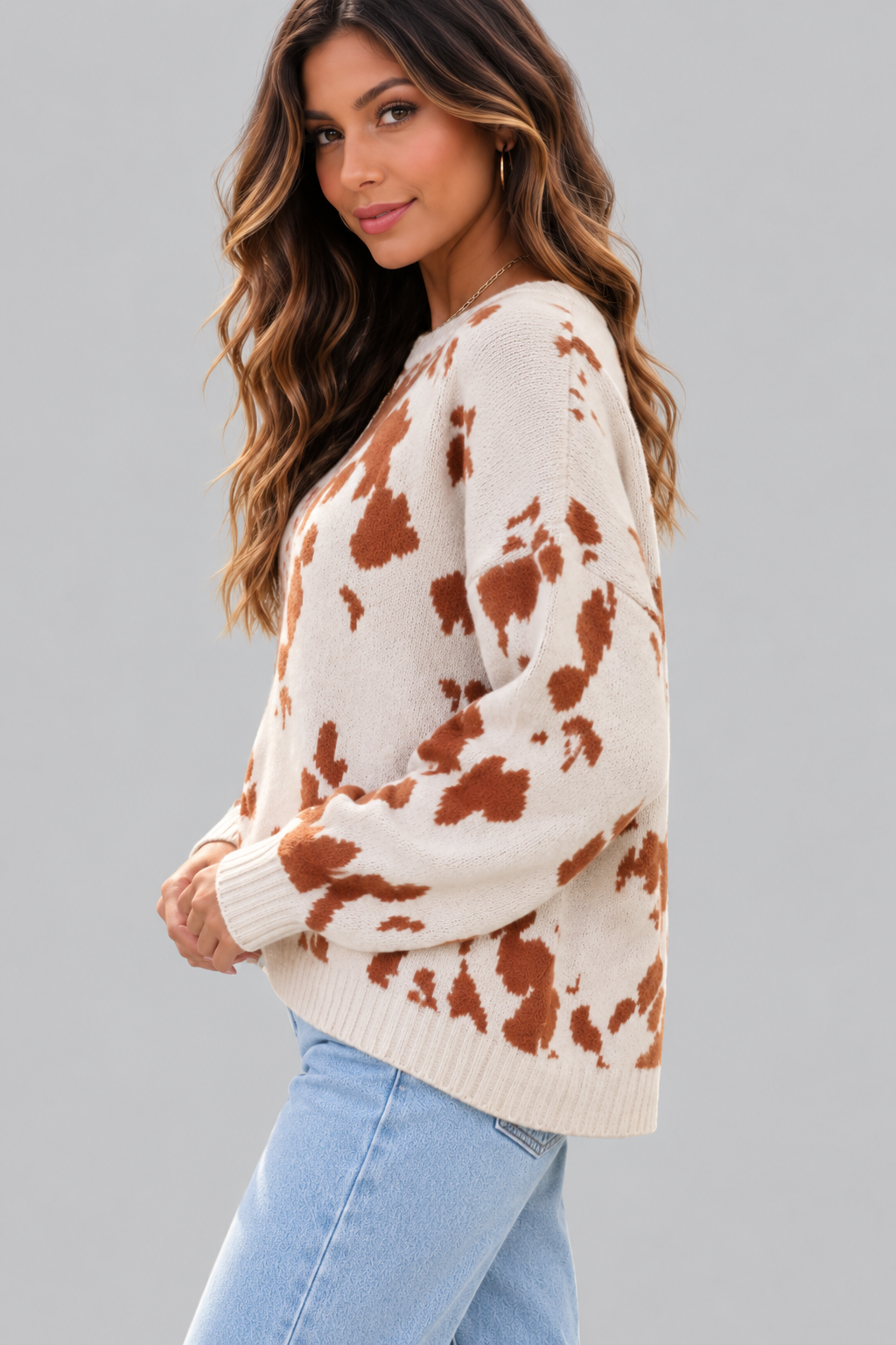 A woman wearing a cream-colored knit sweater with a rust-brown cow-print pattern, paired with light blue jeans, photographed against a solid gray studio background.
