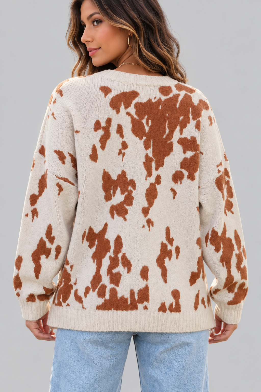 A woman wearing a cream-colored knit sweater with a rust-brown cow-print pattern, shown from the back against a solid gray studio background.