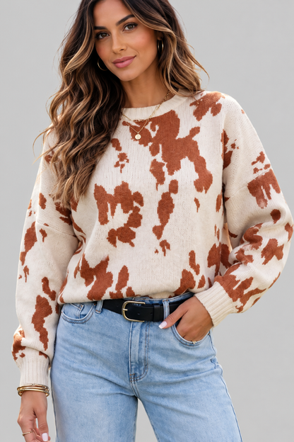 A woman wearing a cream-colored knit sweater with a brown cow-print pattern, paired with light-wash blue jeans and a black belt, against a solid gray studio background.