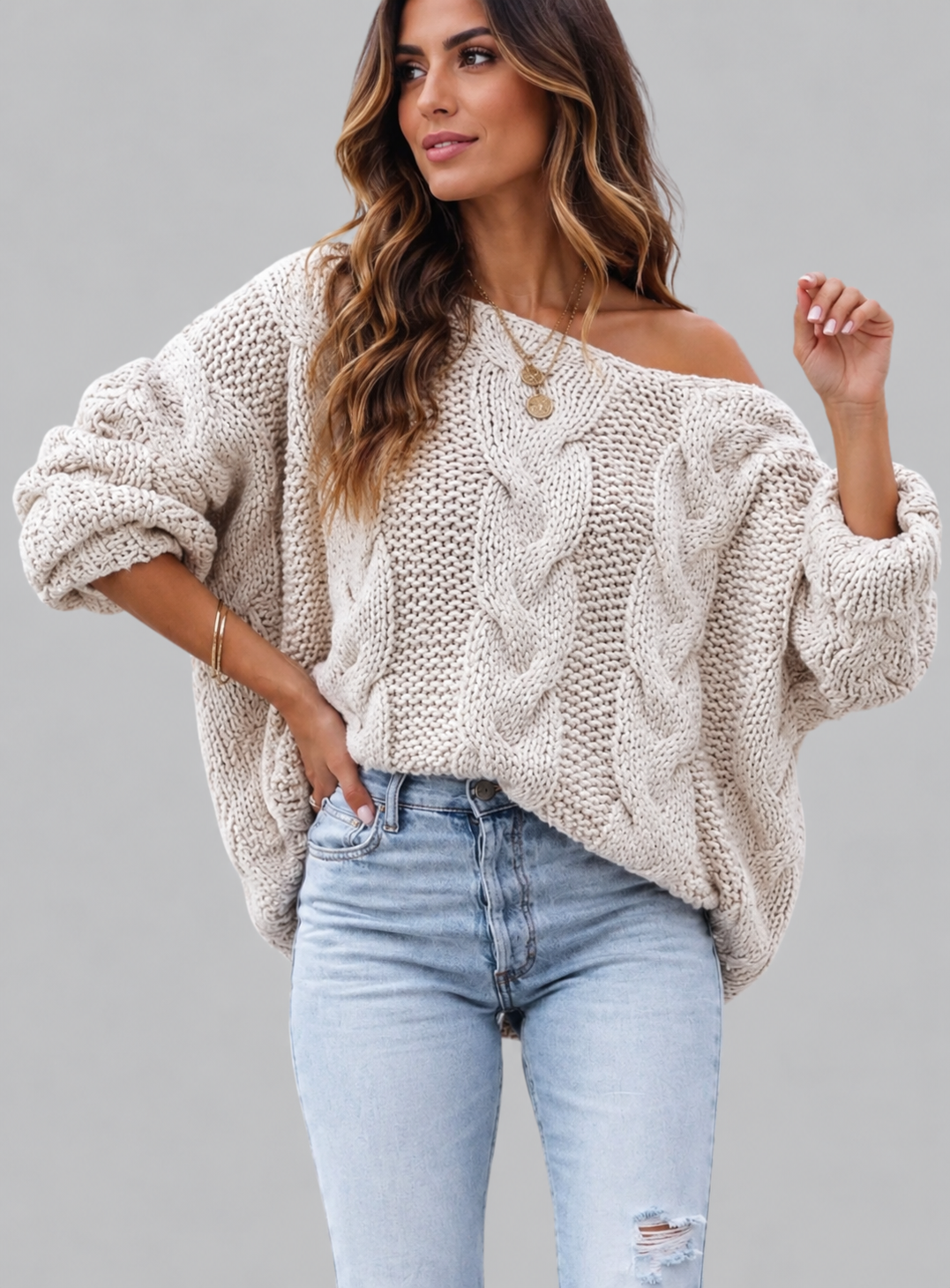 A woman wearing a chunky cable-knit off-the-shoulder sweater in a light oatmeal beige color, paired with light-wash blue jeans, against a solid gray studio background.