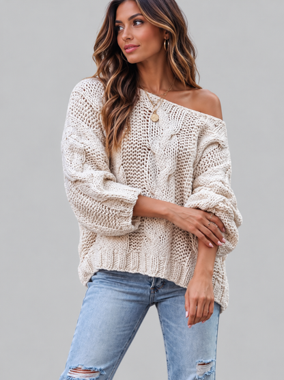 A woman wearing a chunky knit off-the-shoulder sweater in cream and light blue distressed jeans, posed against a solid gray studio background.