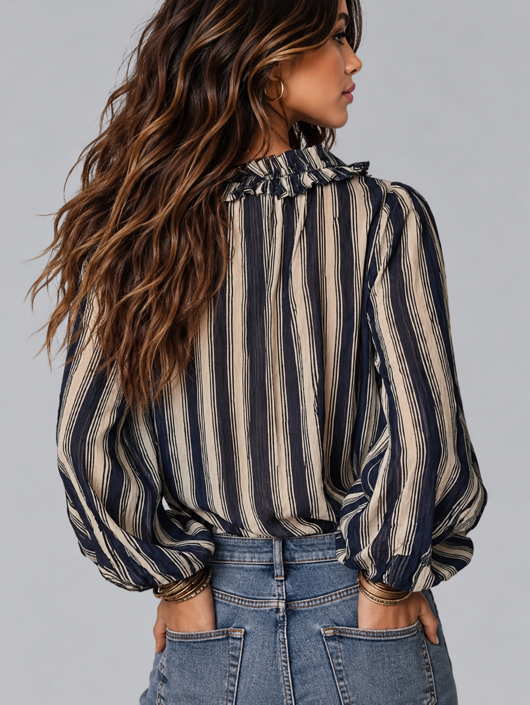 A woman shown from the back wearing a long-sleeve striped blouse with a ruffled high neck and voluminous balloon sleeves, paired with blue jeans against a solid gray studio background.