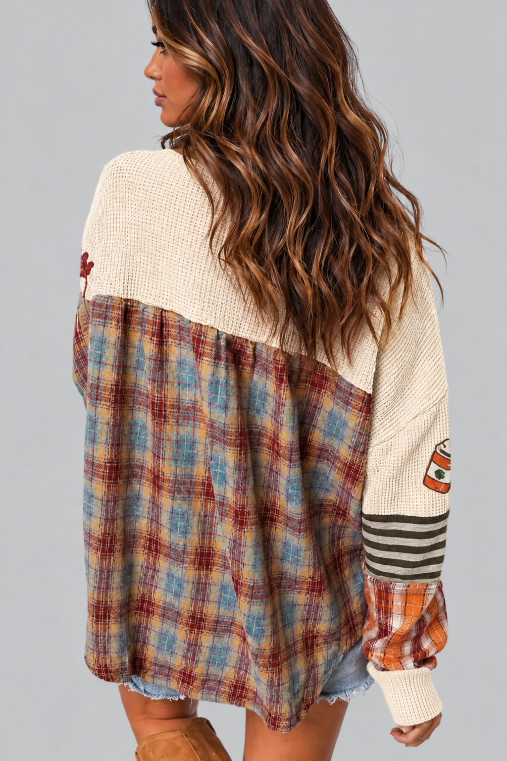 A woman shown from the back wearing a color-block knit sweater with a cream waffle-knit top, a blue and rust plaid middle section, and striped sleeves, photographed against a solid gray studio background.