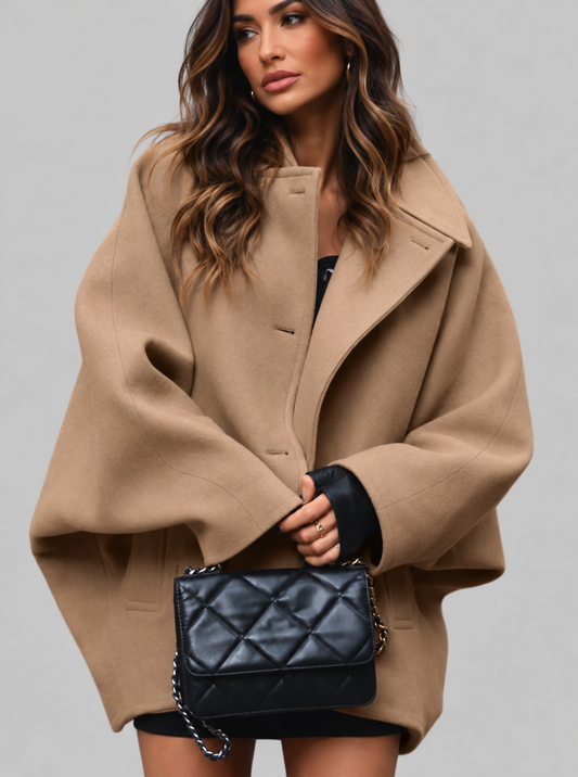 A woman wearing a camel-colored oversized cape coat and holding a black quilted shoulder bag with a silver chain strap, set against a solid gray studio background.
