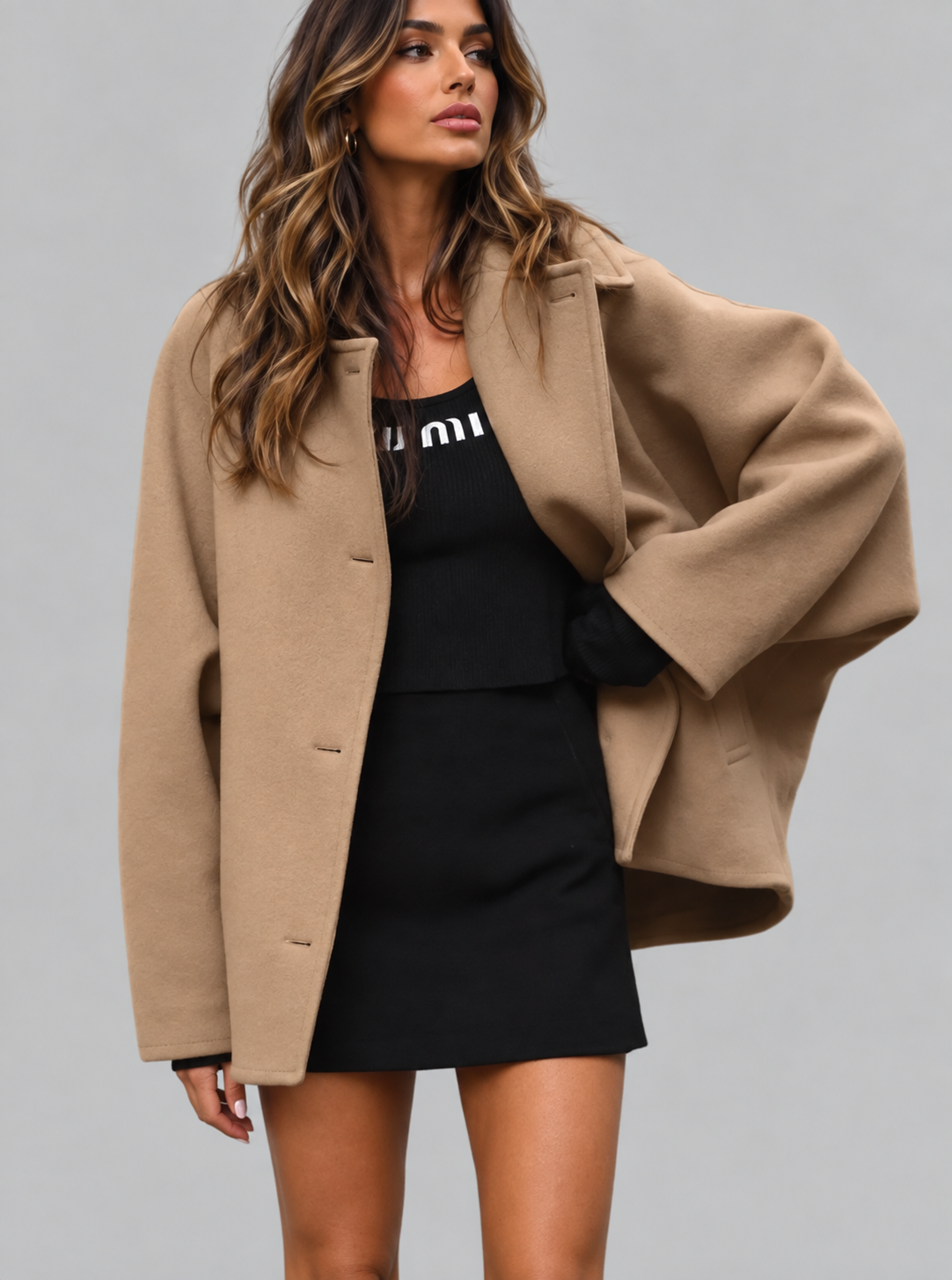 A woman modeling a camel-colored oversized coat over a black dress against a solid gray studio background.