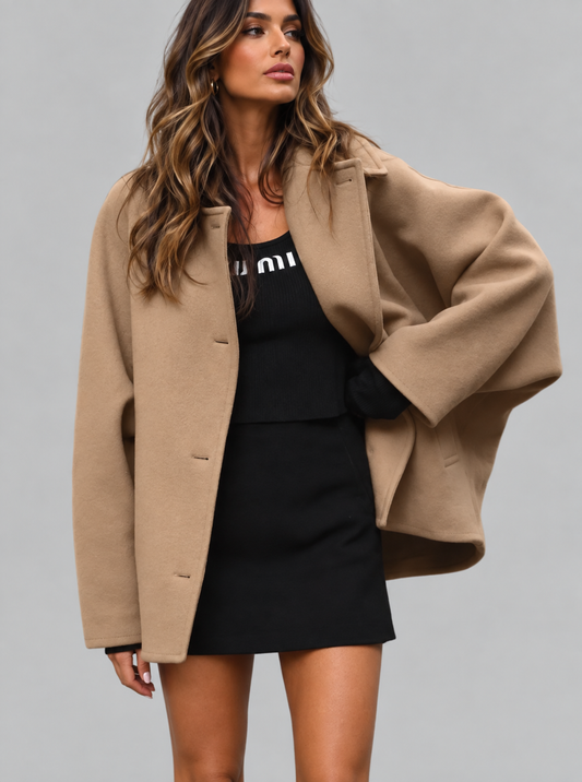 A woman modeling a camel-colored oversized coat over a black dress against a solid gray studio background.