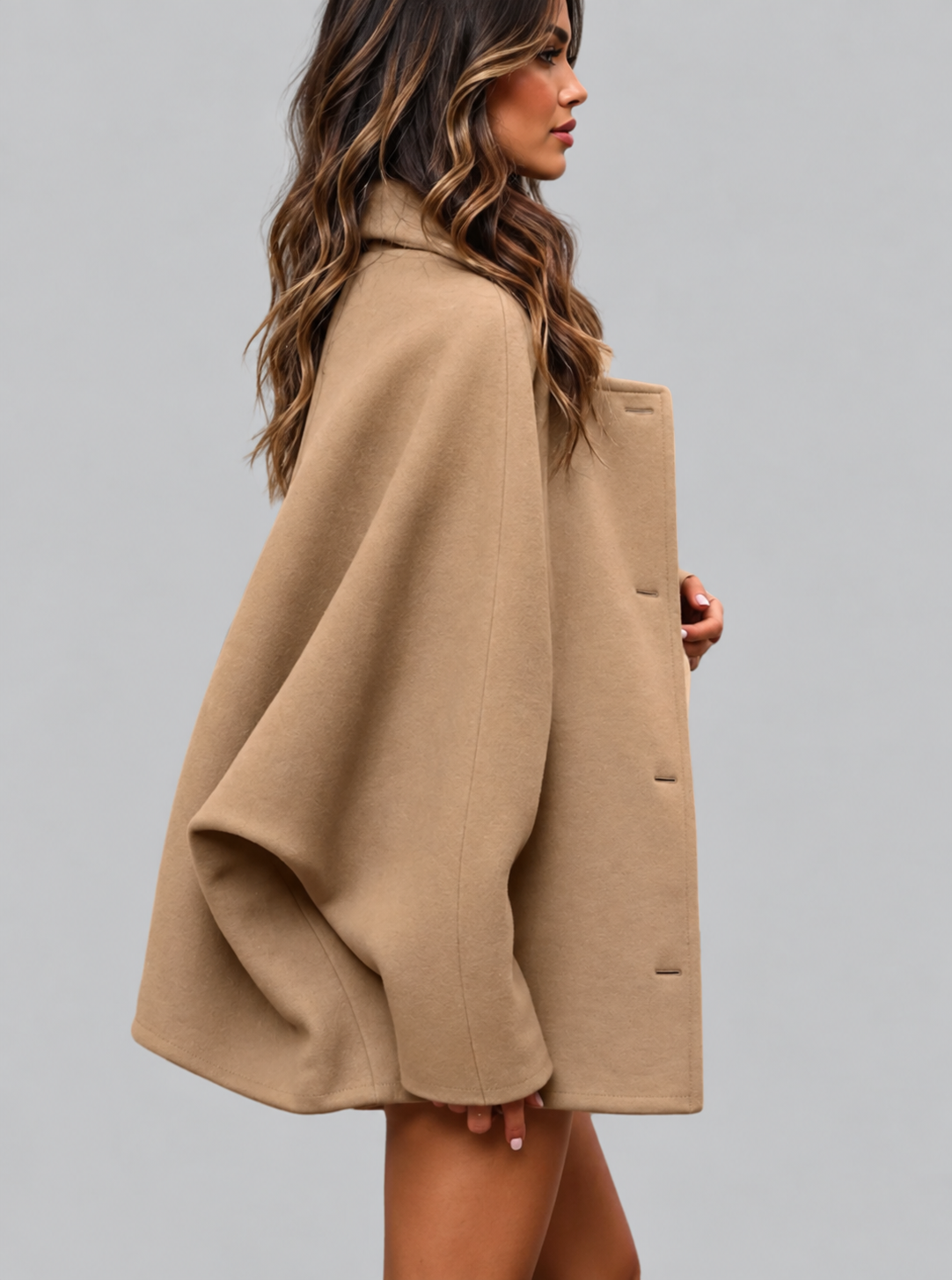 A woman wearing a camel-colored, oversized cape-style coat, shown from a side profile against a solid gray studio background.