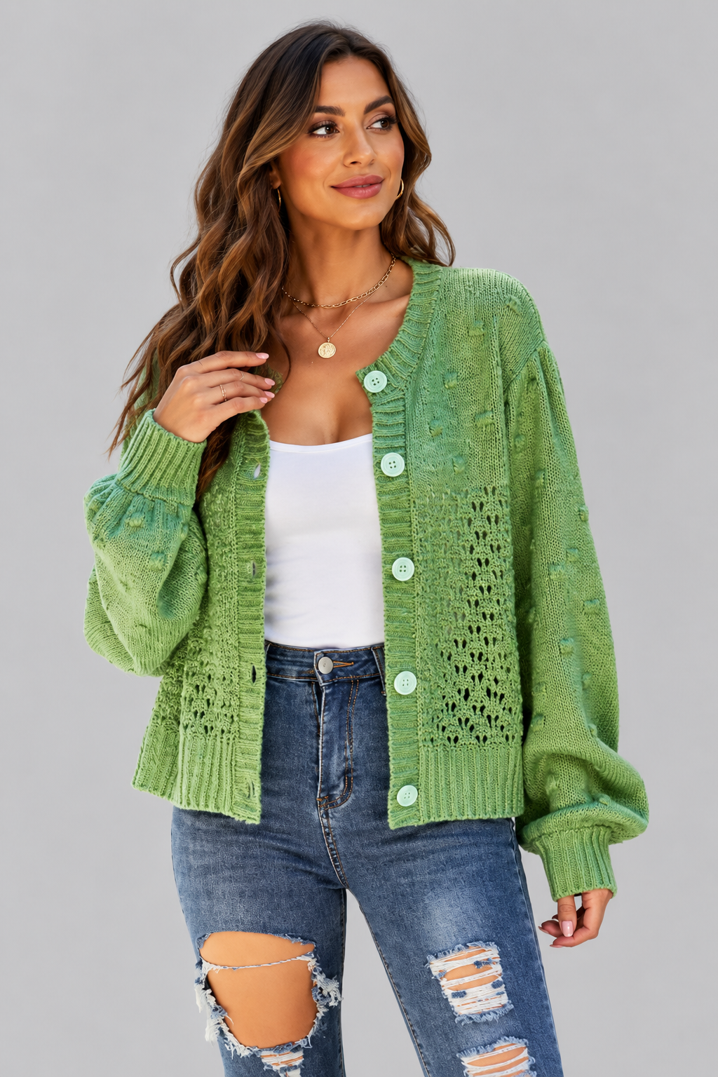 A woman wearing a light green textured knit cardigan with light green buttons, styled over a white tank top and distressed blue jeans, against a solid gray studio background.
