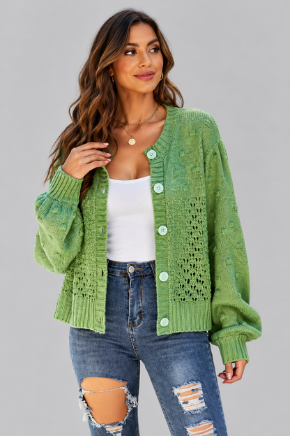 A woman wearing a light green textured knit cardigan with light green buttons, styled over a white tank top and distressed blue jeans, against a solid gray studio background.