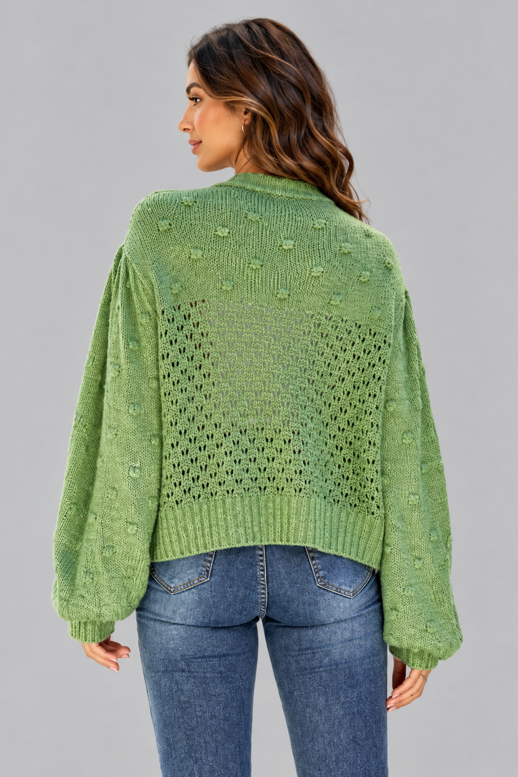 A woman shown from the back wearing a light green knit sweater with textured bobble details and balloon sleeves, paired with blue jeans, against a solid gray studio background.