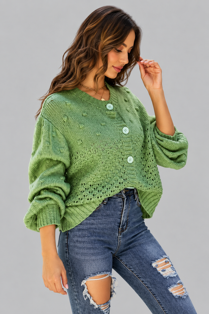A woman wearing a sage green knit button-up sweater with textured bobble details and light green buttons, paired with distressed blue jeans, against a solid gray studio background.