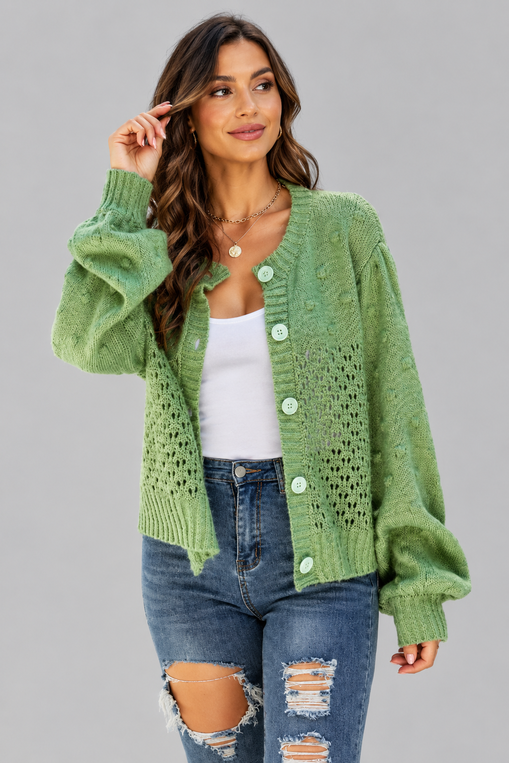 A woman modeling a light green open-front knit cardigan with large buttons, worn over a white tank top and distressed blue jeans against a solid gray studio background.
