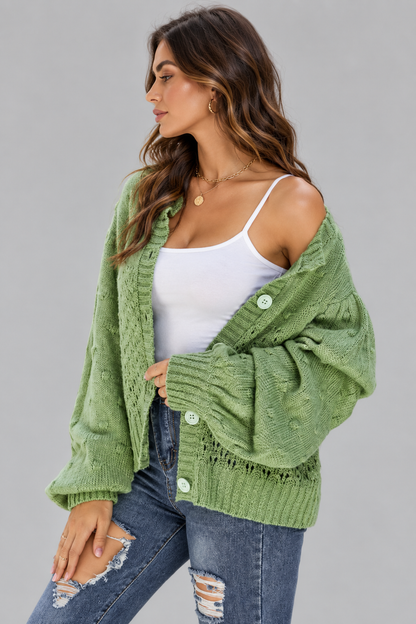 A woman wearing a sage green cable-knit cardigan over a white camisole and distressed blue jeans, posed against a solid gray studio background.