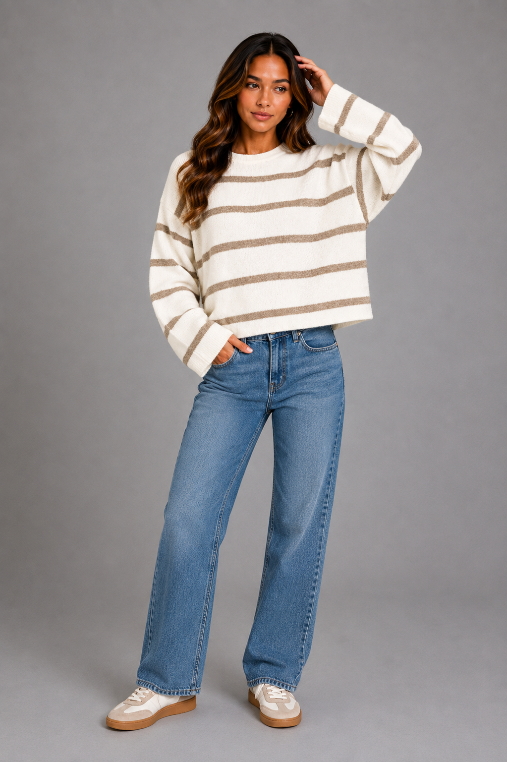 A woman modeling a cream and tan horizontally striped knit sweater, paired with blue jeans and white sneakers, against a solid gray studio background.