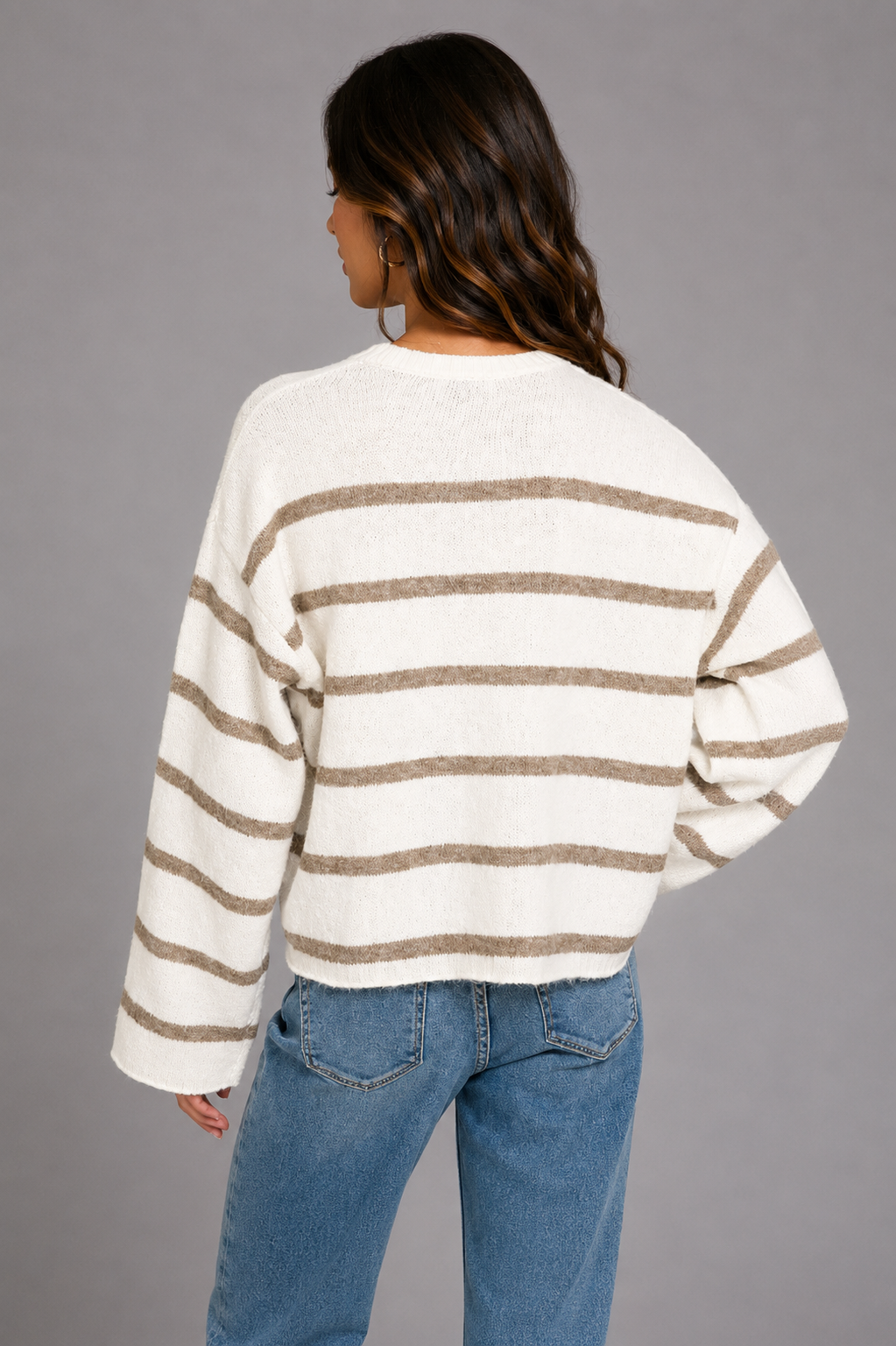 A woman shown from the back wearing a cream-colored knit sweater with horizontal taupe-brown stripes and blue jeans, set against a solid gray studio background.