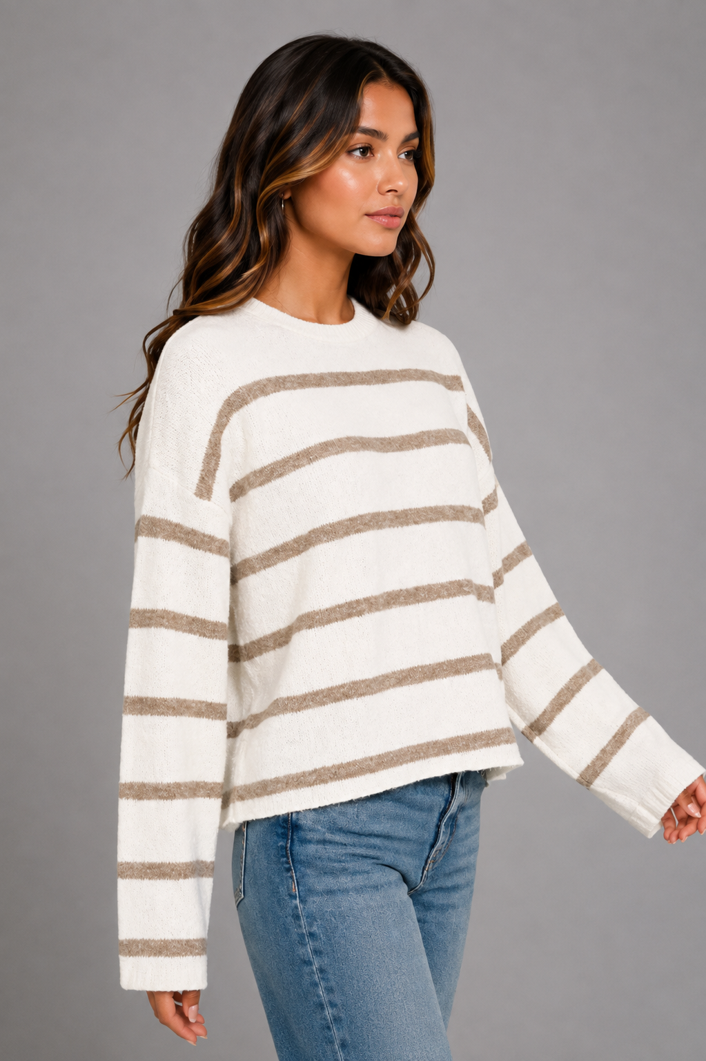 A woman wearing a cream and taupe striped knit sweater with blue jeans, photographed against a solid gray studio background.