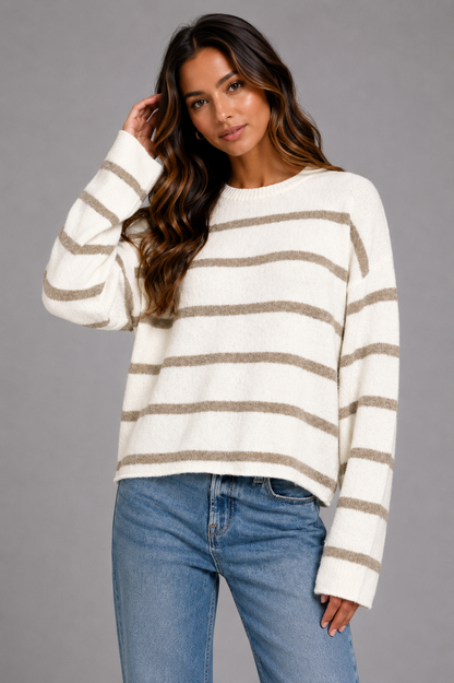 A woman modeling a cream and taupe striped knit sweater with blue jeans against a solid gray studio background.