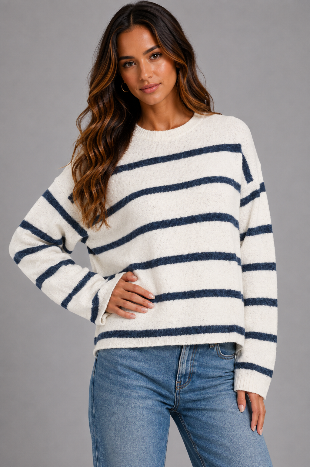 A woman wearing a fuzzy knit crewneck sweater with navy blue horizontal stripes on an off-white background, paired with blue jeans, against a solid gray studio background.
